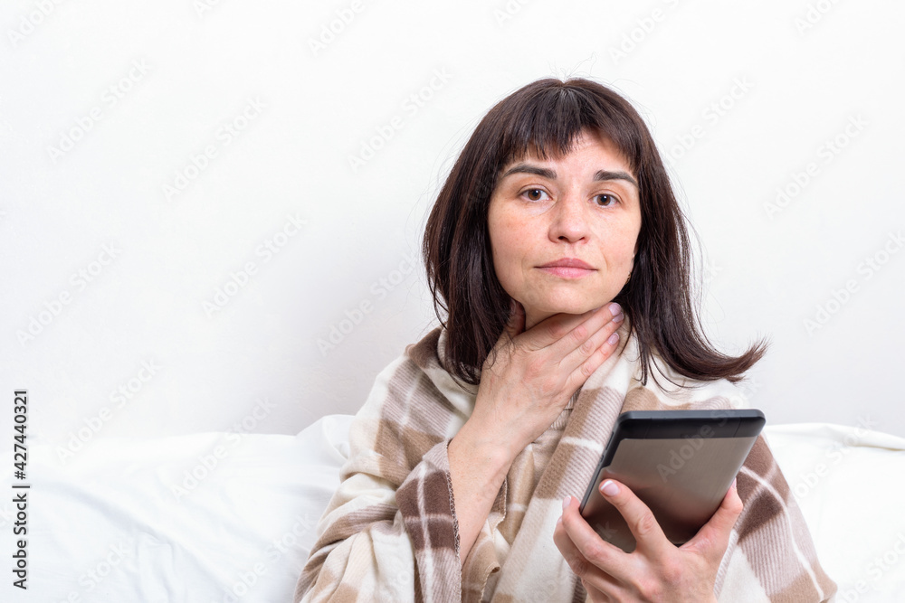 A sick woman covered with a blanket communicating with a doctor on a tablet and showing a sore throat. A sick woman covered with a blanket communicating with a doctor on a tablet and showing a sore throat.