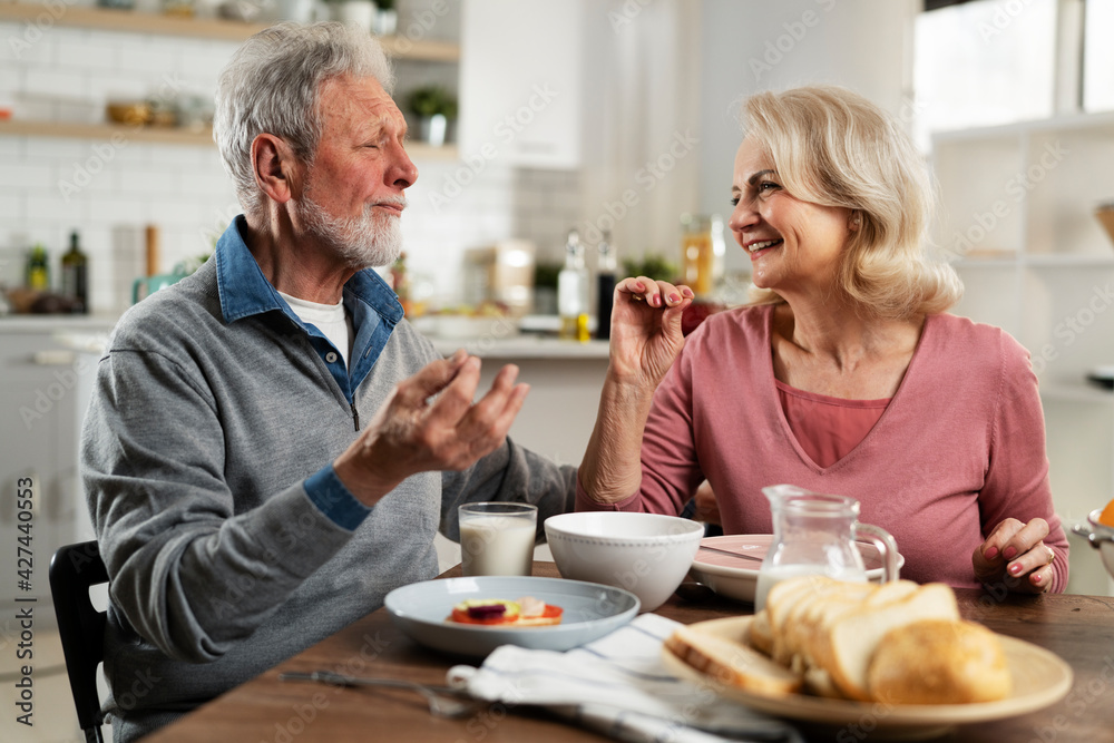 Senior couple eating breakfast in the kitchen. Husband and wife talking and laughing while eating a sandwich. Senior couple eating breakfast in the kitchen. Husband and wife talking and laughing while eating a sandwich.