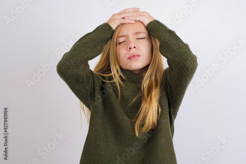 Caucasian kid girl wearing green knitted sweater against white wall suffering from strong headache desperate and stressed because of overwork. Depression and pain concept.