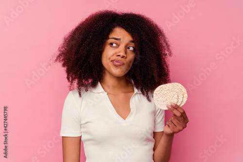 Young african american woman eating a rice cakes isolated on pink background confused, feels doubtful and unsure.