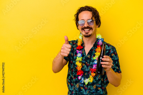 Young caucasian man wearing a hawaiian necklace holding a beer isolated on yellow background smiling and raising thumb up