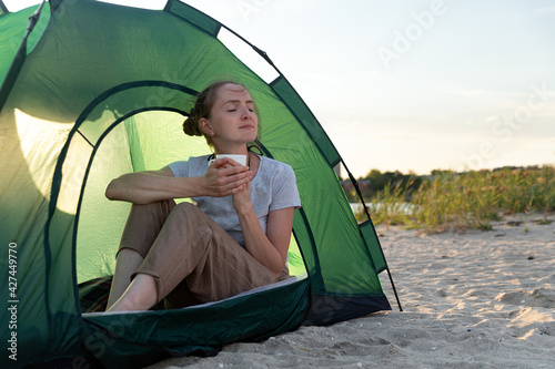 Female tourist sitting in touristic tent with cup of hot tea on sandy beach. Camping