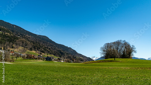 Blick von Düns durch das Großwalsertal zur markanten, verschneiten Berg "Rote Wand". Frühling im Vorarlberg mit Sonnenschein und Schneeresten auf den Bergen. Wald an den steilen Hängen, Wiesen am Dorf