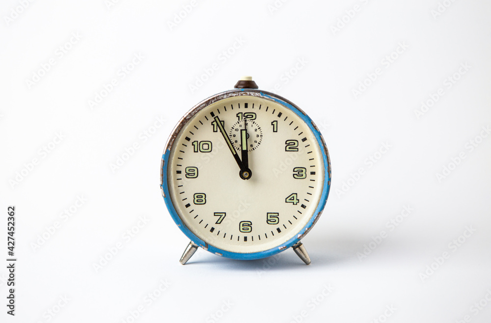 Old analog blue clock on a white background. The clock shows the time from five minutes to twelve o'clock. Old analog blue clock on a white background. The clock shows the time from five minutes to twelve o'clock.