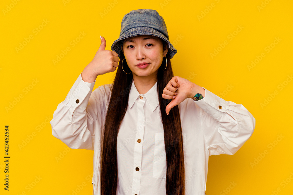 Young chinese woman isolated on yellow background showing thumbs up and thumbs down, difficult choose concept Young chinese woman isolated on yellow background showing thumbs up and thumbs down, difficult choose concept
