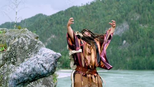 Man in the role of an Altai shaman in national dress with a wolf skin on his shoulder with long hair dances and waves his arms. Shooting a summer day on a bear-shaped mountain and a fast-flowing river