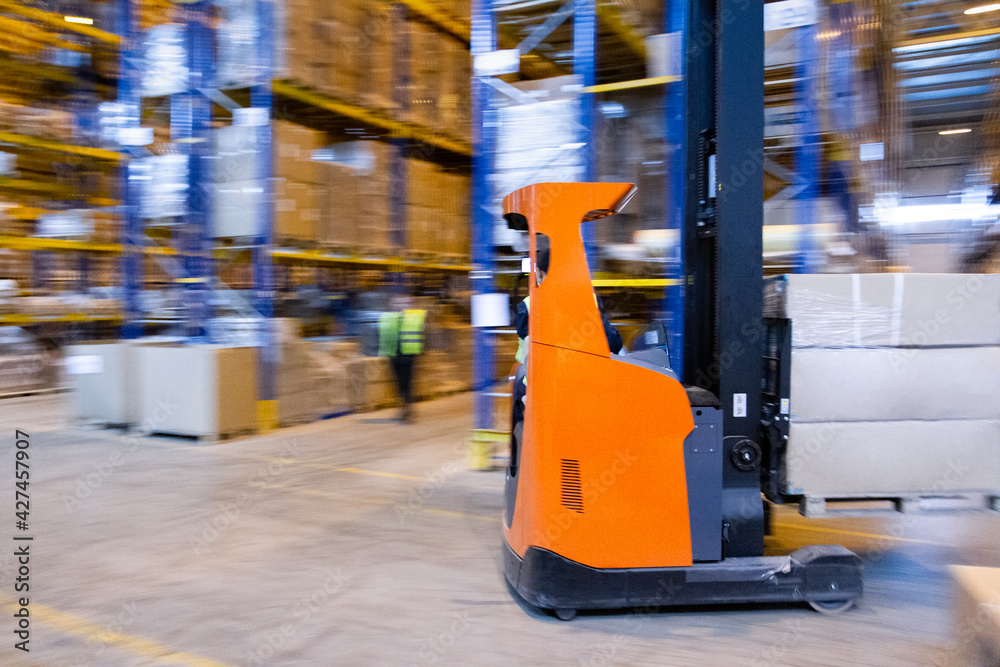Fototapeta premium Warehouse worker driving a forklift truck full of goods between high blue yellow steel shelves inside warehouse