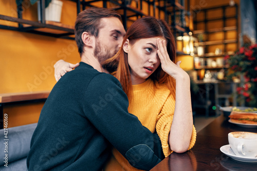a woman with disgust hugs a man in a sweater at a table in a cafe