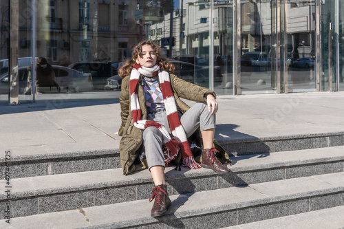 A teenage student Caucasian girl with curly brown hair in casual spring clothes sits on the steps of a modern building with a glass facade
