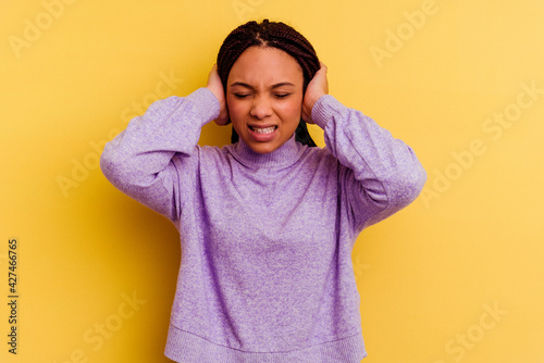 Young african american woman isolated on yellow background covering ears with hands trying not to hear too loud sound. © Asier