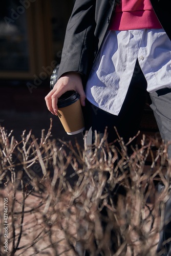 A woman in a black men's suit and white shirt with a disposable cup of coffee in her hands on a sunny day.