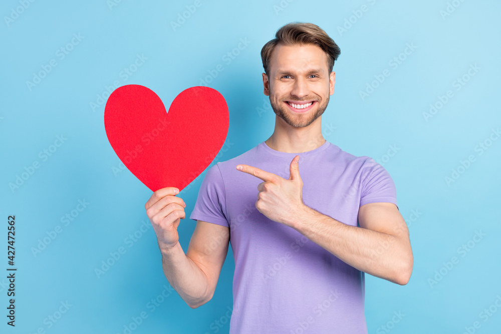 Portrait of cheerful guy direct finger paper heart symbol card toothy smile isolated on blue color background Portrait of cheerful guy direct finger paper heart symbol card toothy smile isolated on blue color background