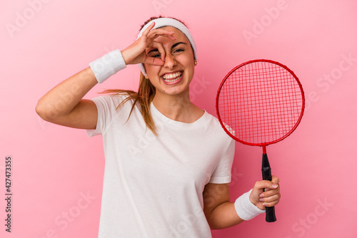 Young cute blonde caucasian woman holding a badminton racket isolated on pink background excited keeping ok gesture on eye.