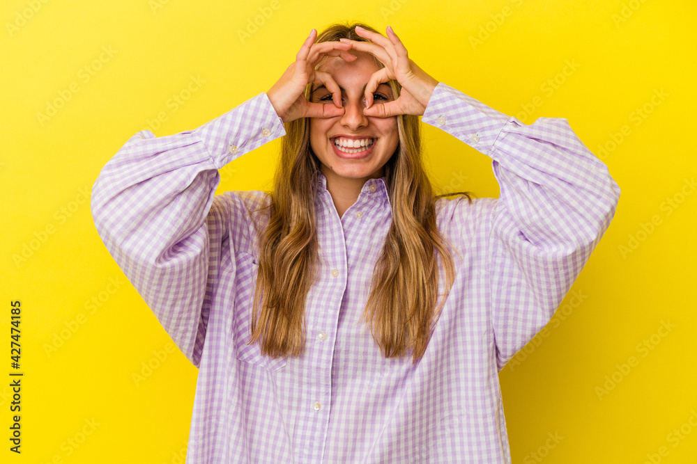 Young blonde caucasian woman isolated on yellow background showing okay sign over eyes