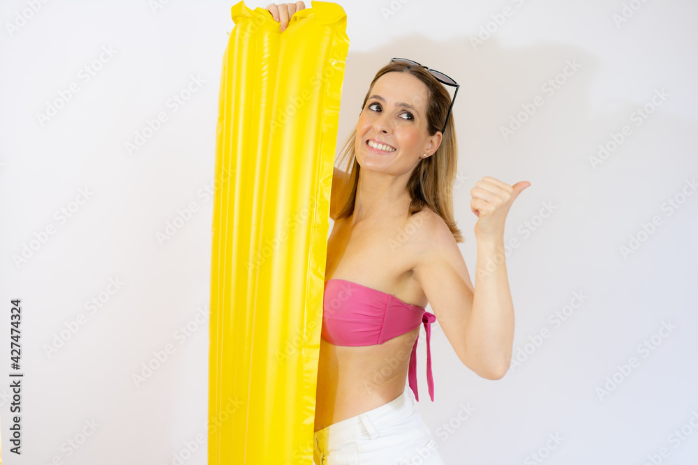 Obraz premium Smiling young woman wearing pink bikini posing with yellow inflatable mattress with thumb up over white background