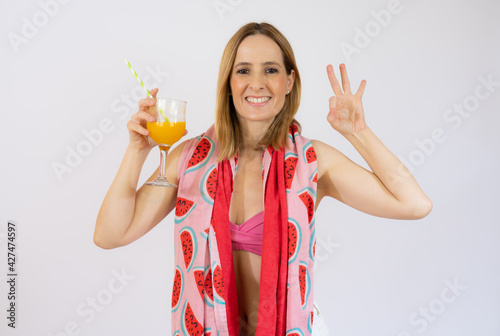 Woman wearing bikini and beach towel holding glass of juice with okay sign