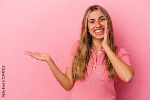 Young blonde caucasian woman isolated on pink background holds copy space on a palm, keep hand over cheek. Amazed and delighted.