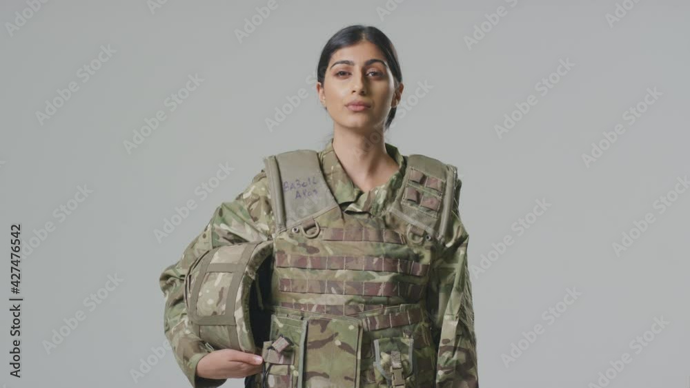 Young female soldier in uniform wearing body armour and holding helmet in front of plain studio background - shot in slow motion