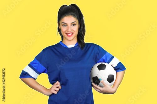 A young soccer player holds her ball under her arm in front of a yellow background. © Simone