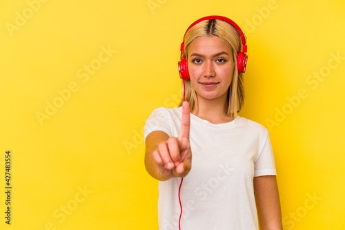 Young venezuelan woman listening music isolated on yellow background showing number one with finger.