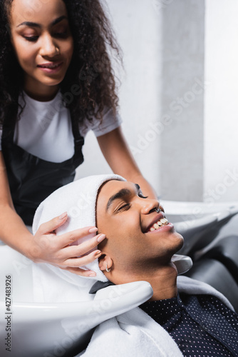 Young african american client smiling near sink and hairdresser with towel on blurred background