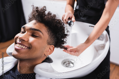 Positive african american man sitting near sink and blurred hairdresser