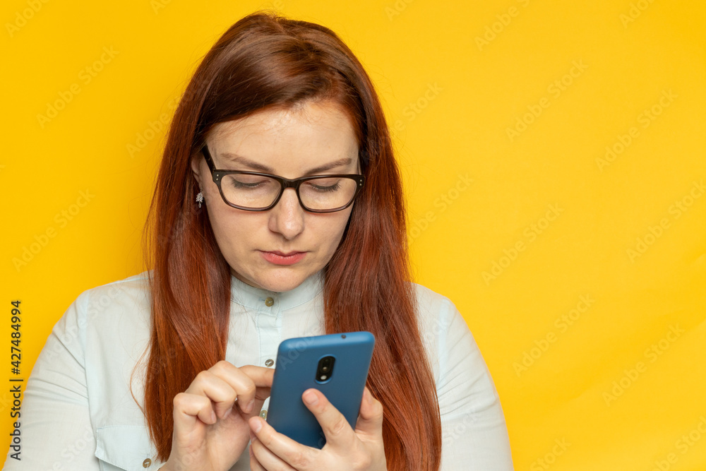 Focused business woman uses smartphone while standing against a yellow wall background Focused business woman uses smartphone while standing against a yellow wall background