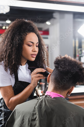 Focused african american hairstylist trimming hair of client in cape on blurred foreground