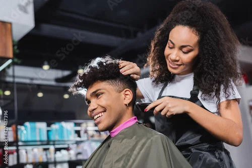 Fototapeta na wymiar Smiling african american hairdresser in apron trimming neck of client in salon