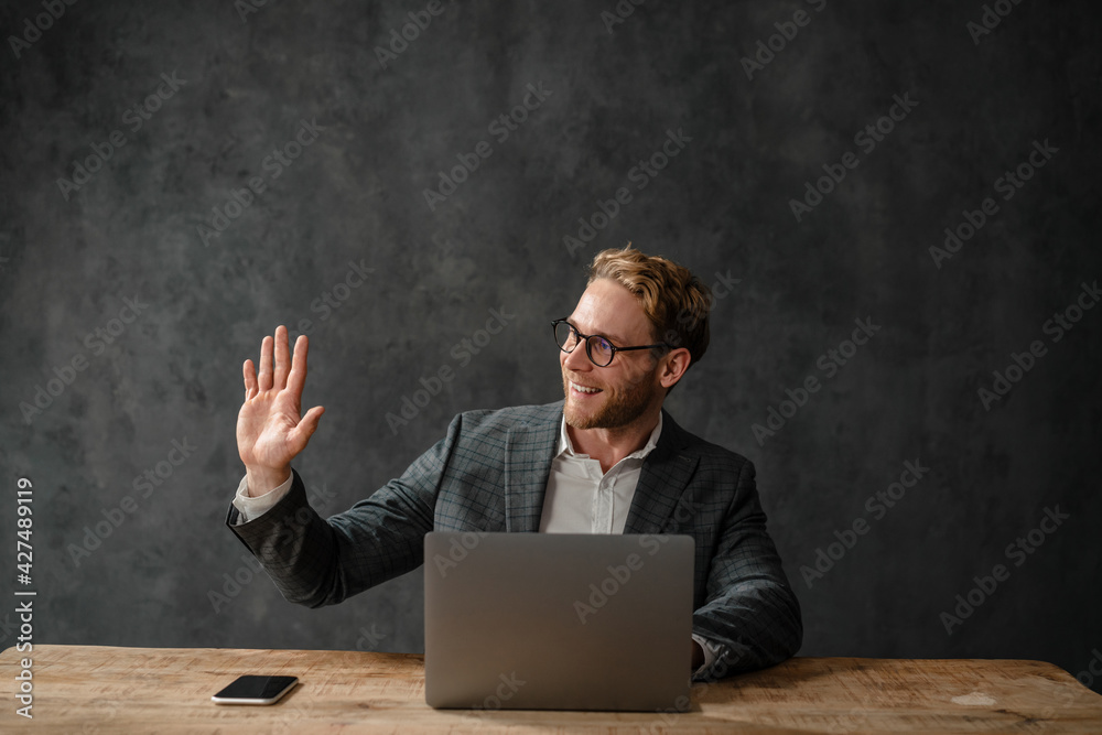 The smiling man waving his hand to the side while sitting in front of a laptop at a table in the studio The smiling man waving his hand to the side while sitting in front of a laptop at a table in the studio