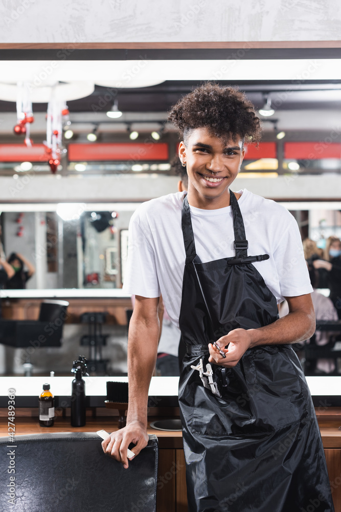 Smiling african american hairdresser in apron holding comb and scissors