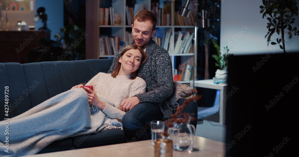 Charming young caucasian couple of married attractive man and woman staying home lying on sofa watching favorite comedy movie on TV.