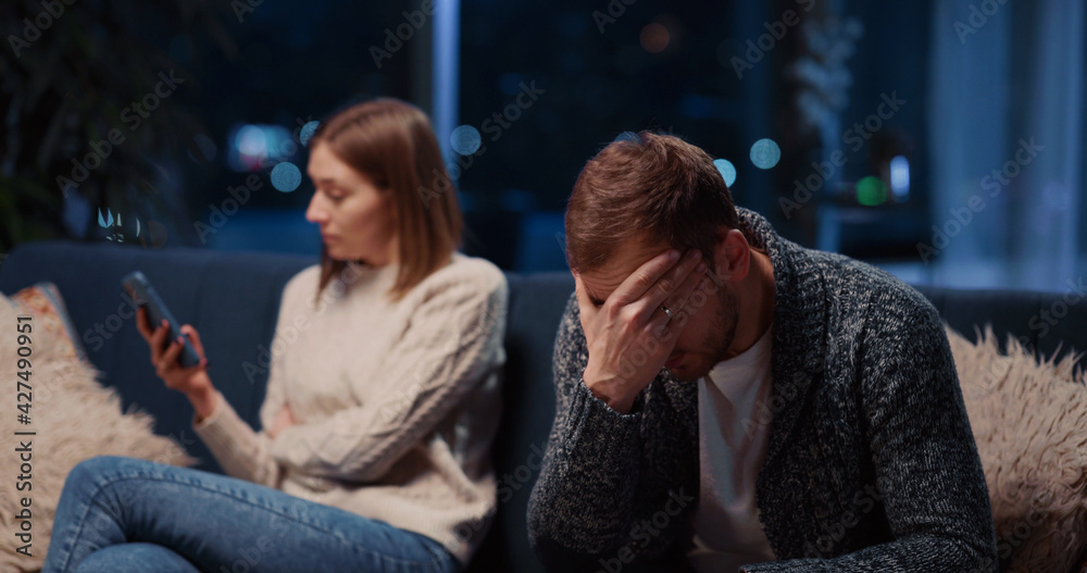 Caucasian young married couple making up conflict in their apartment. Angry young woman fell jealous about her husband reading messages on his smartphone. Caucasian young married couple making up conflict in their apartment. Angry young woman fell jealous about her husband reading messages on his smartphone.