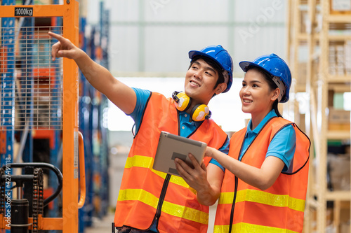Group of Asian young male and female employee warehouse worker in safety vest and helmet working with tablet for checking products or parcel goods on shelf pallet in industry factory warehouse