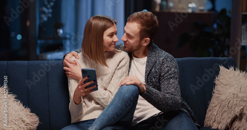 Charming caucasian couple staying home using mobile phone together. Affectionate young happy girlfriend and boyfirend dating in apartment at night. photo