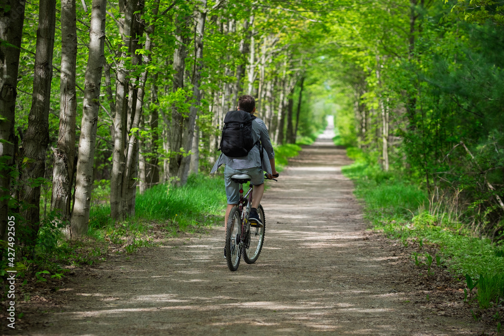 Young man biking cycling through the park alley green tunnel made of tree brunches. Summer, spring scene. Recreational sport and cycling concept. Selective focus. Caledon trailway path, Ontario. Young man biking cycling through the park alley green tunnel made of tree brunches. Summer, spring scene. Recreational sport and cycling concept. Selective focus. Caledon trailway path, Ontario.