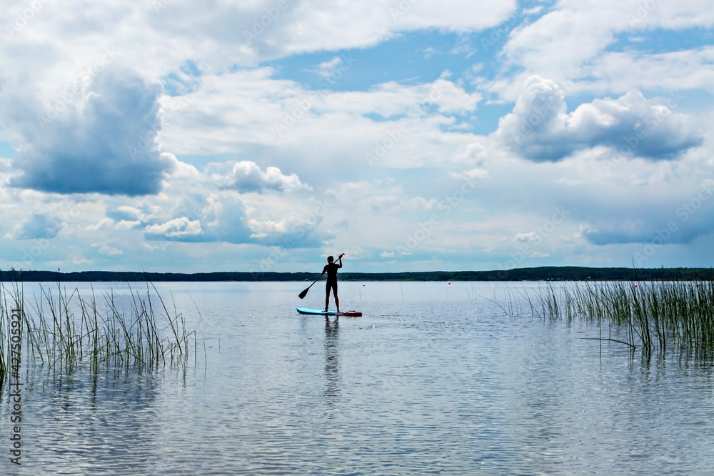 young man from the back in black thermal suit paddleboarding on blue stand up paddle board on lake against background of cloudy sky, male silhouette, active lifestyle, outdoor activities, sup