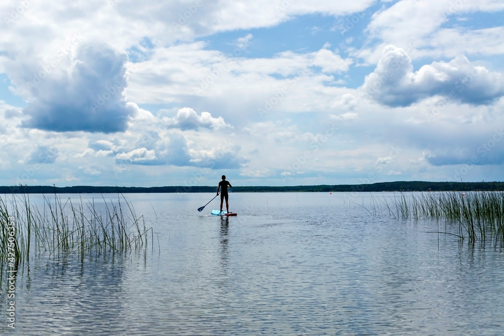 young man from the back in black thermal suit paddleboarding on blue stand up paddle board on lake against background of cloudy sky