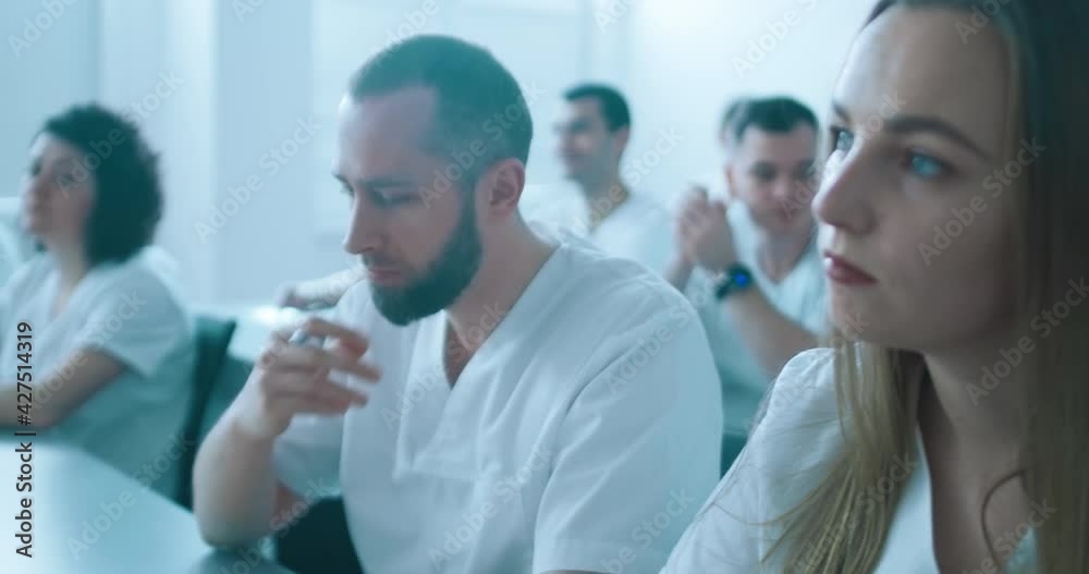 Close-up a female medical worker and male medical worker with the beard who is sitting in the desk and listening somebody in the lecture hall with other medical workers. Close-up a female medical worker and male medical worker with the beard who is sitting in the desk and listening somebody in the lecture hall with other medical workers.