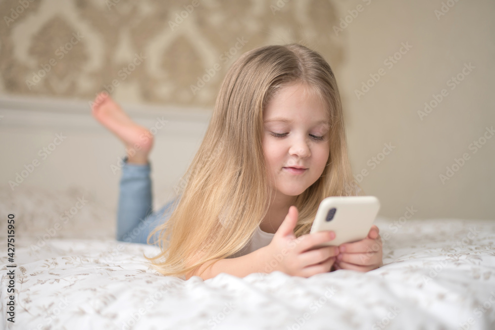 A little girl lies on the bed and plays on the phone social internet.