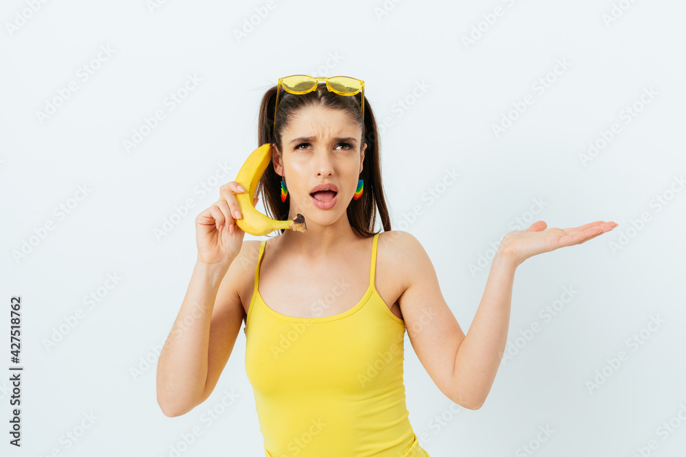 Displeased young woman in summer outfit and yellow sunglasses holds banana as phone Displeased young woman in summer outfit and yellow sunglasses holds banana as phone