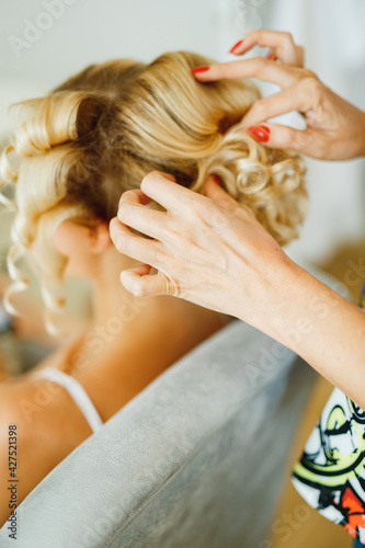 Stylist doing hair to blonde bride during preparation for wedding ceremony, close-up 
