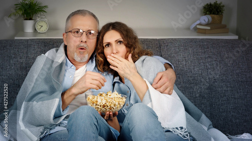 A middle-aged couple watches horror movies on TV and eats popcorn on the couch.