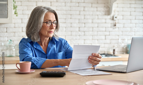 Adult senior 60s woman working at home at laptop. Serious middle aged woman at table holding document calculating bank loan payments, taxes, fees, retirement finances online with computer technologies photo