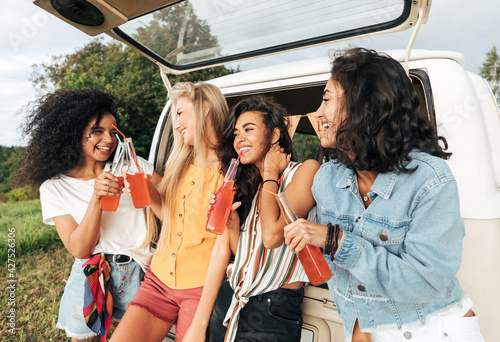 Group of diverse female friends toasting bottles near camper van. Four women enjoying road trip.