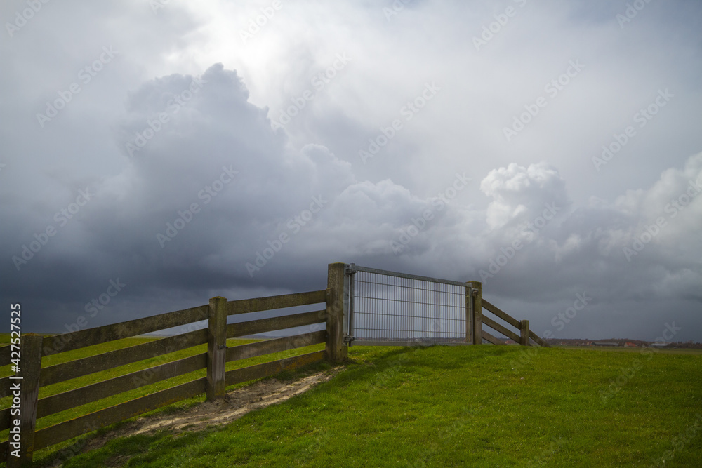 Dutch dike with grass cover, a fence made of planks and steel on a grassy embankment under dark and threatening storm clouds