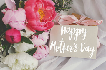 Happy mother's day. Happy mother's day text on card and  beautiful peonies bouquet, gift box on soft white fabric. Stylish floral greeting card. Handwritten lettering. Mothers day