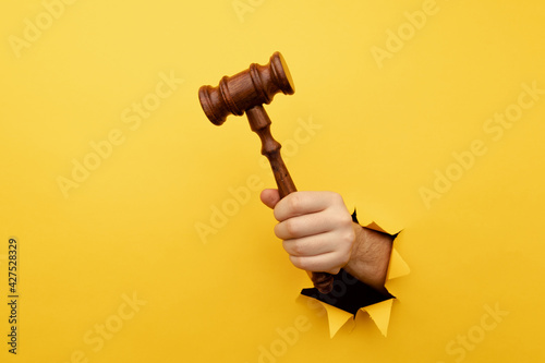 Hand holding a judge's gavel through torn yellow paper wall. Law and justice concept