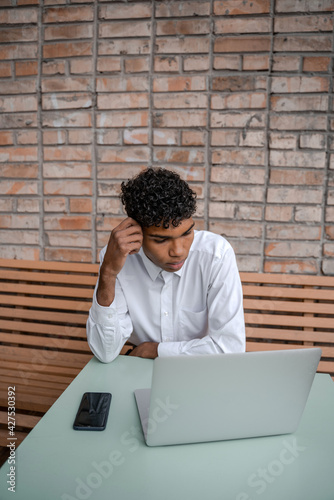 vertical photo A black man sits on a summer terrace or veranda near a cafe. The guy works at a laptop and uses a smartphone. View of the spring business downtown