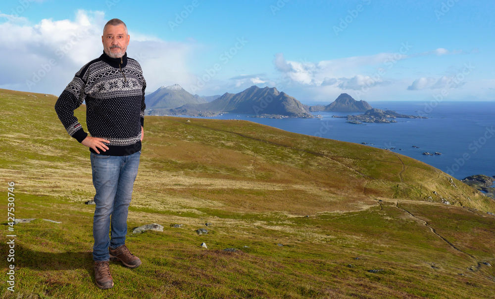 An elderly Norwegian man with a beard is wearing a typical sweater and is standing on a mountain. The Lofoten Islands can be seen in the background.